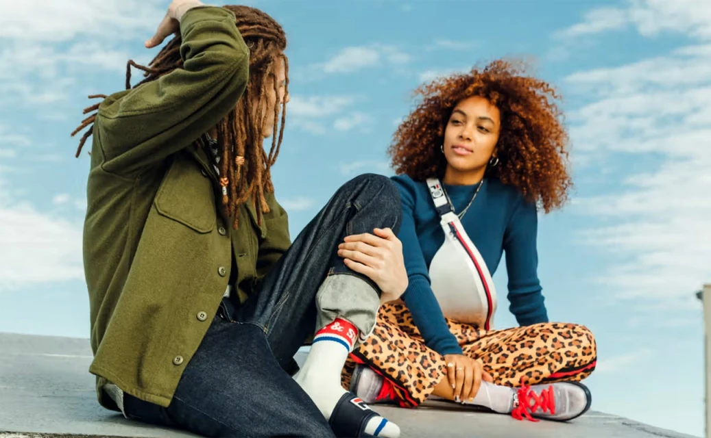 Two young adults with dreadlocks and curly red hair are sitting outside in the sunshine on a rooftop, smiling and talking to each other.