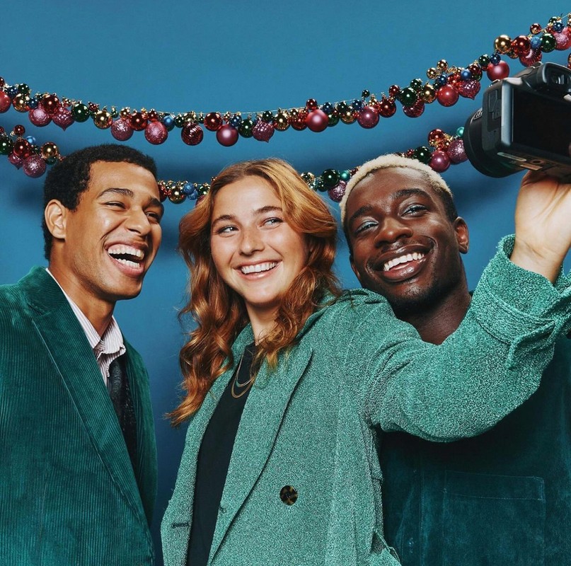 Three happy people in stylish teal corduroy clothing smiling at the camera held by one of them, indoors with festive Christmas ornaments.