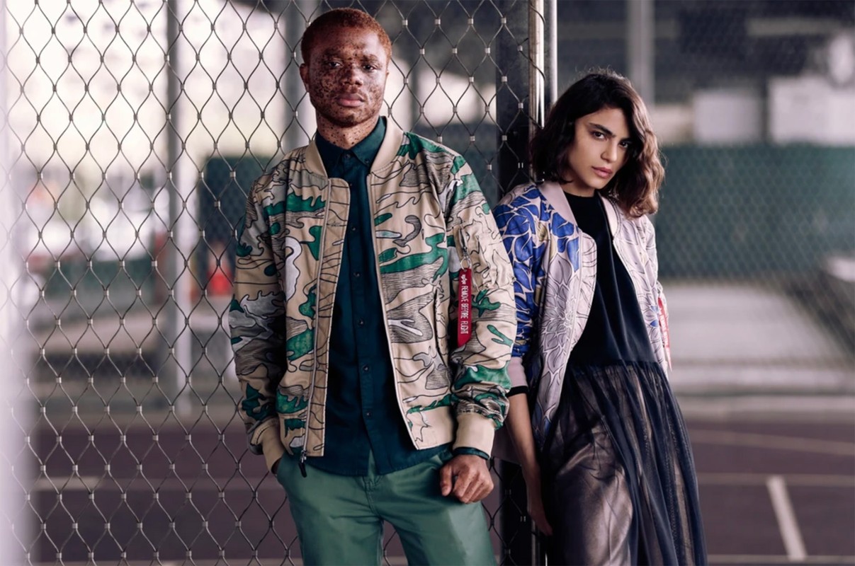 Two fashion models pose in patterned jackets against a wire fence, with a soft focus emphasizing their modern, street-style apparel.
