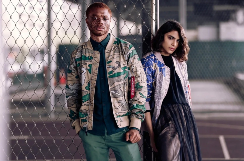 Two fashion models pose in patterned jackets against a wire fence, with a soft focus emphasizing their modern, street-style apparel.