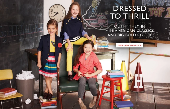 Three young girls in fashionable outfits pose in a classroom setting with a chalkboard, books, and school-related props, smiling at the camera.
