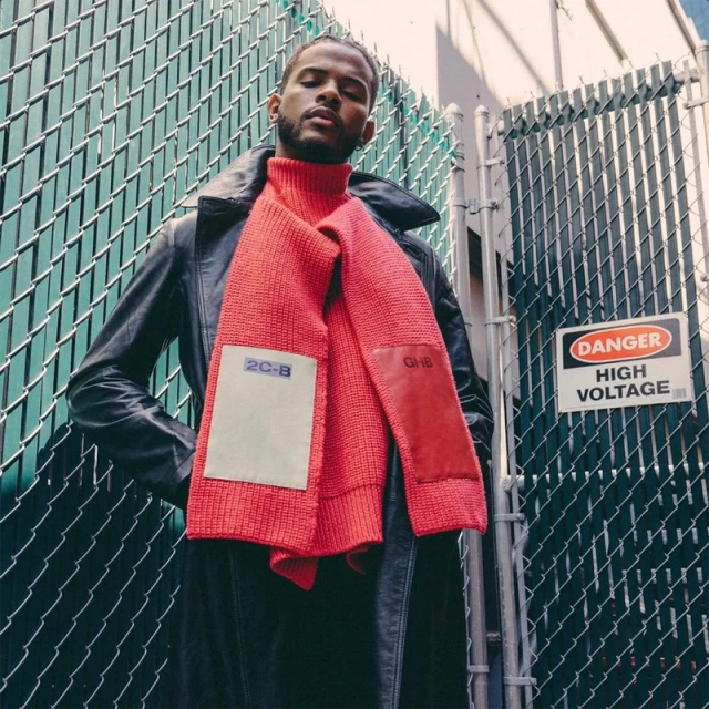 Adult male in outdoor fashion shot wears black coat and red scarf with chain-link fence and danger sign background in medium shot.