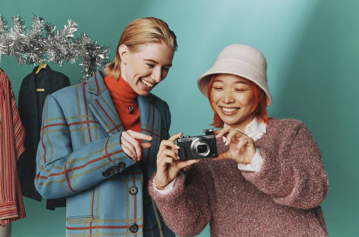Two women smiling together in front of a light blue background, one holding a camera; silver tinsel and hanging clothes in the background.