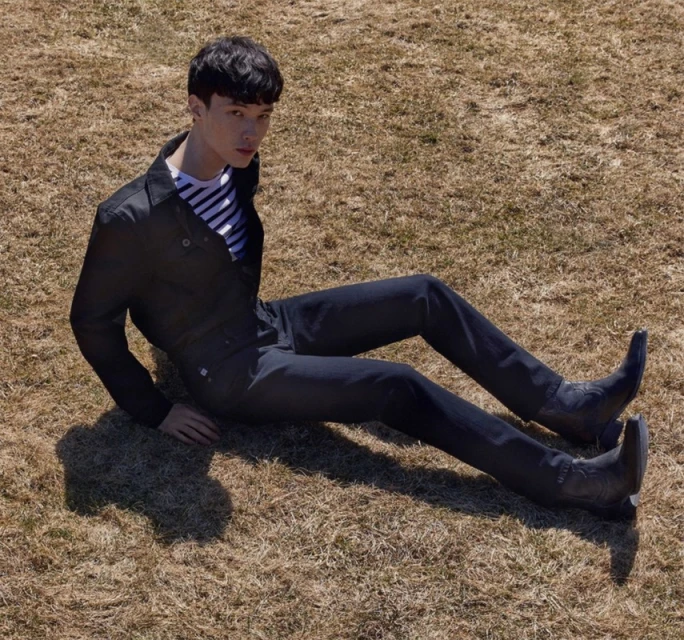 Man sitting on dry grass in black jacket and jeans. He wears striped shirt and cowboy boots. Bright sunlight and outdoor setting.