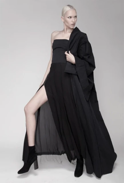 Fashion shot of a slender female model in a strapless black top, long black skirt with thigh-high slit, and black jacket on grey background.