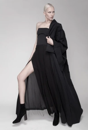 Fashion shot of a slender female model in a strapless black top, long black skirt with thigh-high slit, and black jacket on grey background.