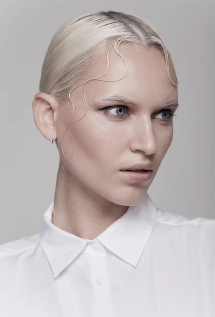 Close-up portrait of a light-skinned woman with short, platinum blonde hair and a white collared shirt against a plain grey background.