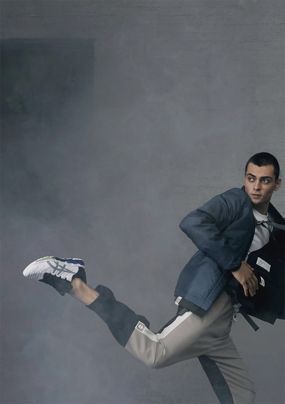 Fashionable adult male in mid-air wearing a blue jacket, white shirt, and track pants, paired with sneakers, posing in front of a grey background.