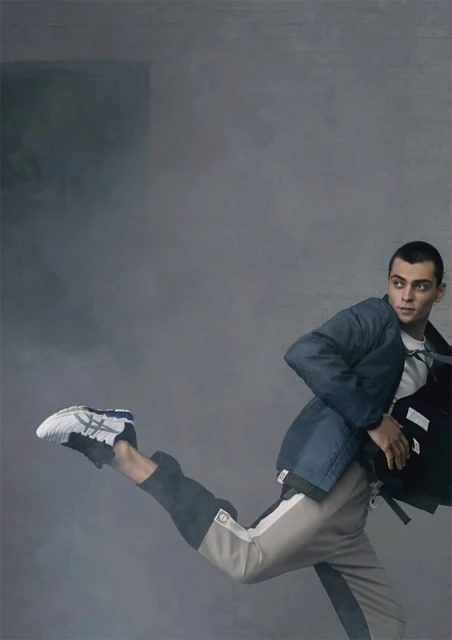 Fashionable adult male in mid-air wearing a blue jacket, white shirt, and track pants, paired with sneakers, posing in front of a grey background.