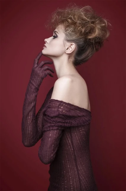 Profile of a woman with stylish updo, wearing long gloves and draped clothing, set against a solid dark red background.