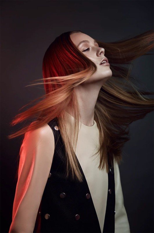 Portrait of a young woman with long, flowing brown hair wearing a black vest and cream-colored top against a dark background.