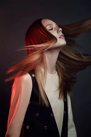 Close-up profile portrait of a woman with long red and blonde hair whipping through the air against a dark grey background.