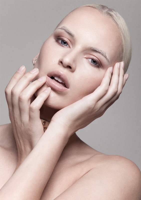Close-up portrait of an adult female with pale skin, blonde hair, light eye makeup, and bare shoulders against a grey studio backdrop.