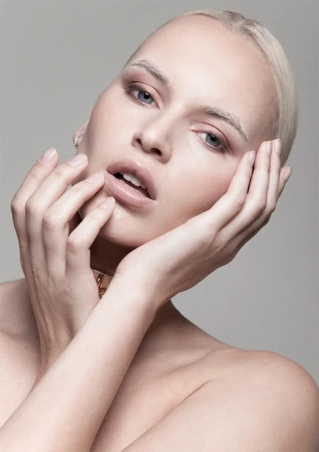 Close-up portrait of an adult female with pale skin, blonde hair, light eye makeup, and bare shoulders against a grey studio backdrop.