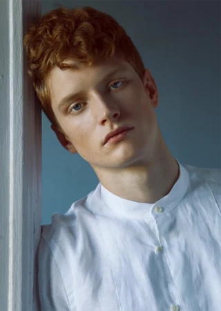 Close-up portrait of a young man with curly red hair wearing a white collared shirt leaning against a wall