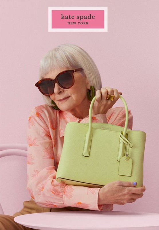 Elderly woman with short white hair posing with a lime green satchel handbag in a pastel pink room for a Kate Spade advertisement.