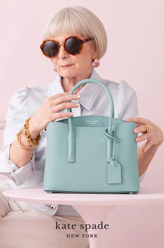 Elderly woman with sunglasses and a light blue Kate Spade purse posing against a light pink background in a studio setting.