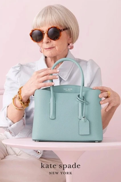 Elderly woman with sunglasses and a light blue Kate Spade purse posing against a light pink background in a studio setting.