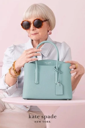 Elderly woman with sunglasses and a light blue Kate Spade purse posing against a light pink background in a studio setting.