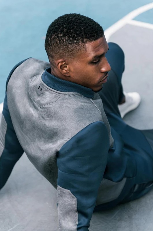An African-American man sits on a blue and grey basketball court in a grey and navy athletic suit, wearing white athletic shoes.