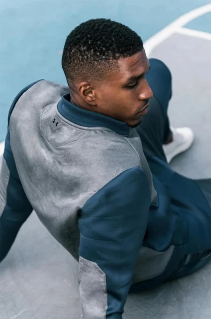 An African-American man sits on a blue and grey basketball court in a grey and navy athletic suit, wearing white athletic shoes.