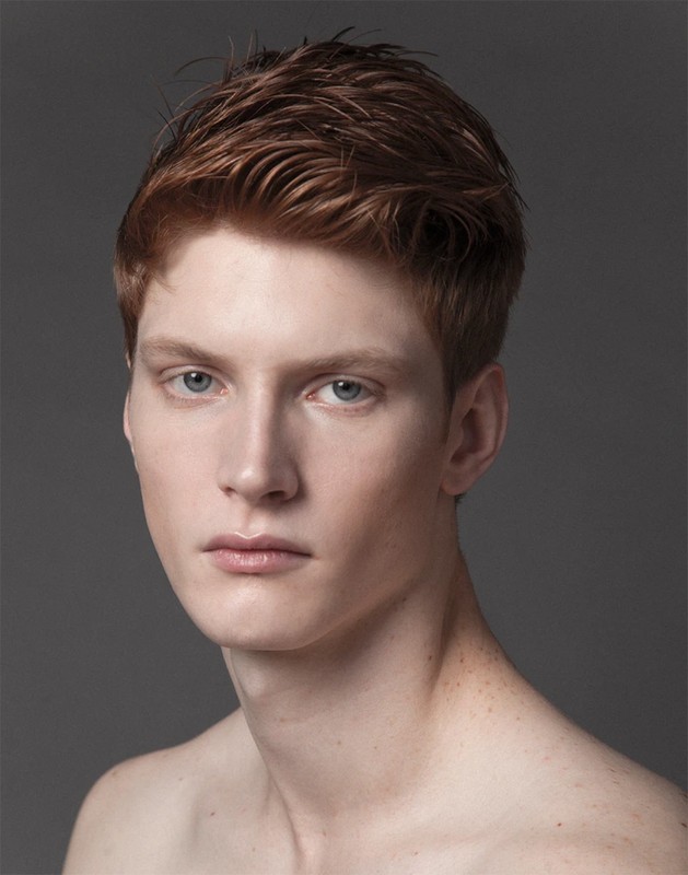Close-up portrait of a young man with short textured reddish-brown hair against a solid grey background