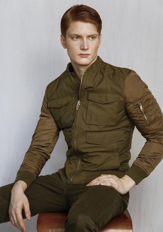 Portrait of a red-haired man in an olive green bomber jacket and pants, seated on a stool against a plain grey background, looking away.