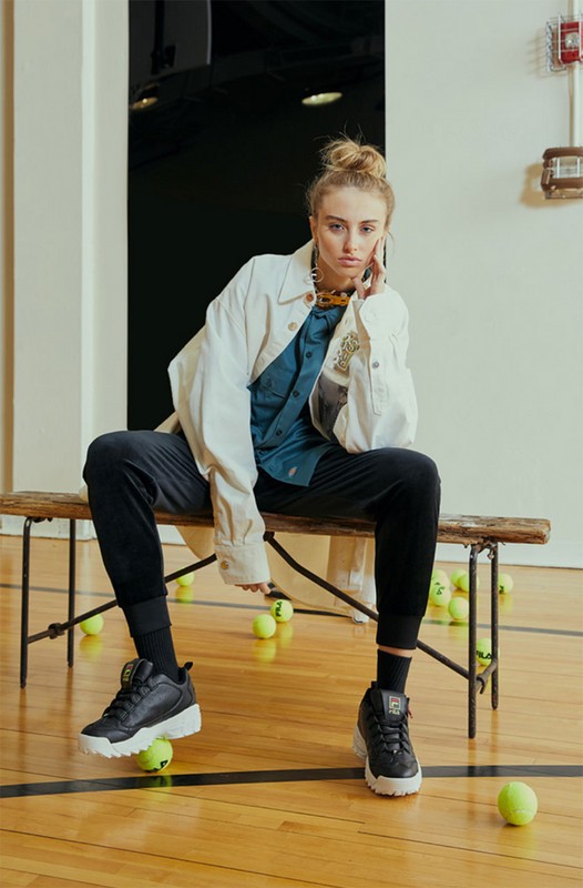 Young woman sits on a bench in an indoor court with tennis balls, wearing a white jacket, teal shirt, black pants, and black and white shoes.