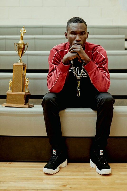 Portrait of a Black man in a red jacket seated on bleachers next to a trophy inside a gymnasium with hardwood floors.