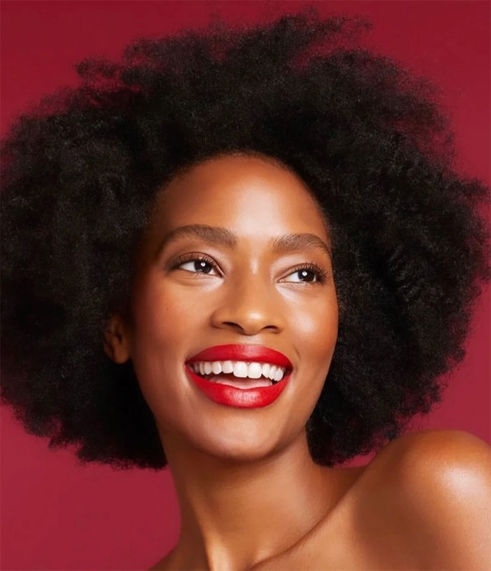 Close-up portrait of a smiling Black woman with voluminous natural afro hair and bold red lipstick against a solid red background