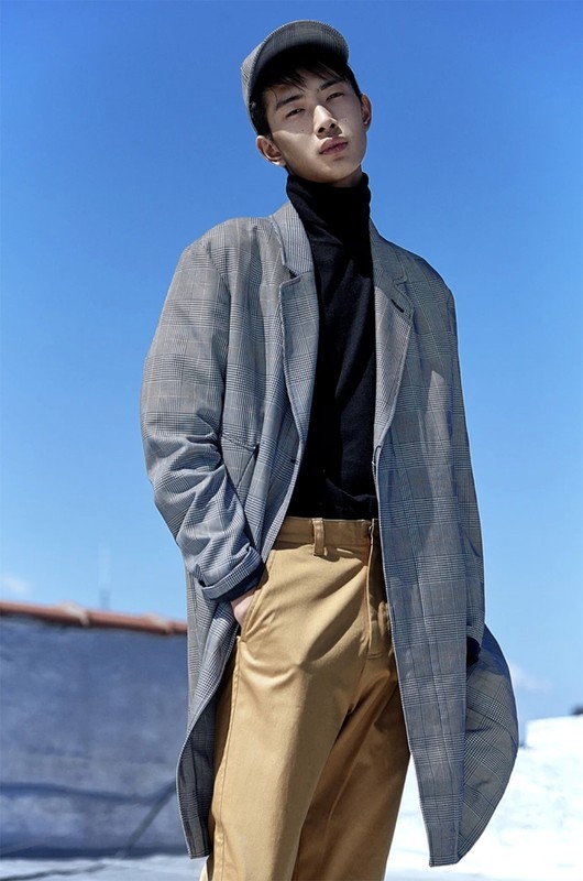 Young man in black turtleneck, khaki pants, and gray plaid coat poses against a bright blue sky in a stylish outdoor fashion portrait.