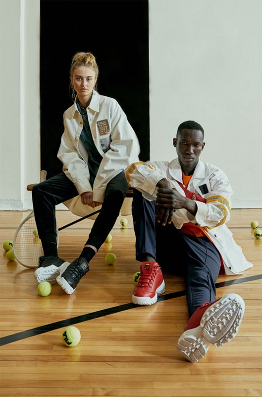 Two models in sportswear sit on a bench surrounded by tennis balls. One is wearing red sneakers, the other black sneakers.