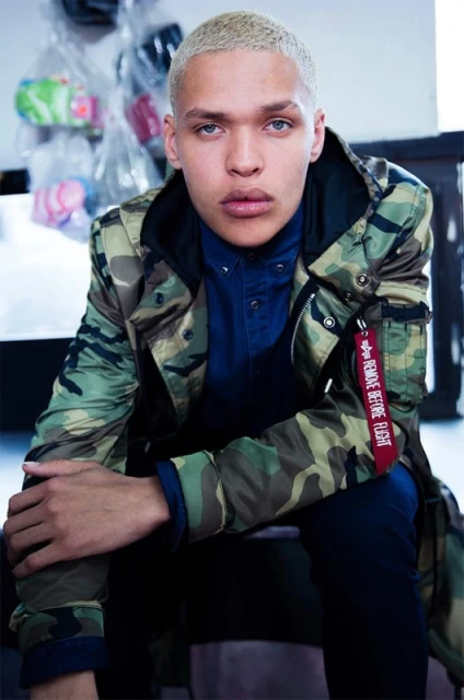 A young adult male with a short, blond buzzcut wears a camouflage jacket and sits indoors looking toward the camera in a medium shot.