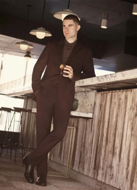 Adult man in a suit leans against a marble bar holding a cocktail in an indoor setting with overhead lighting and bar stools.
