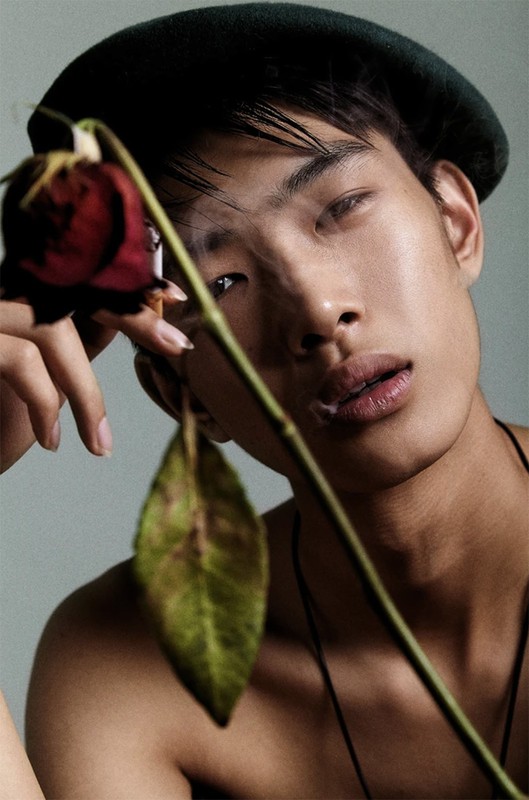 Close-up portrait of a bare-chested Asian male model in a beret, holding a red rose. Studio lighting with a neutral background.