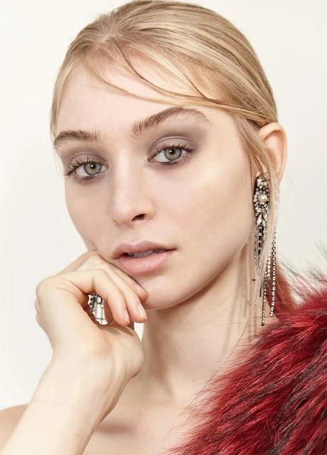 Close-up portrait of a woman with light hair and light skin wearing long, dangling earrings and a red, faux fur stole, indoors.