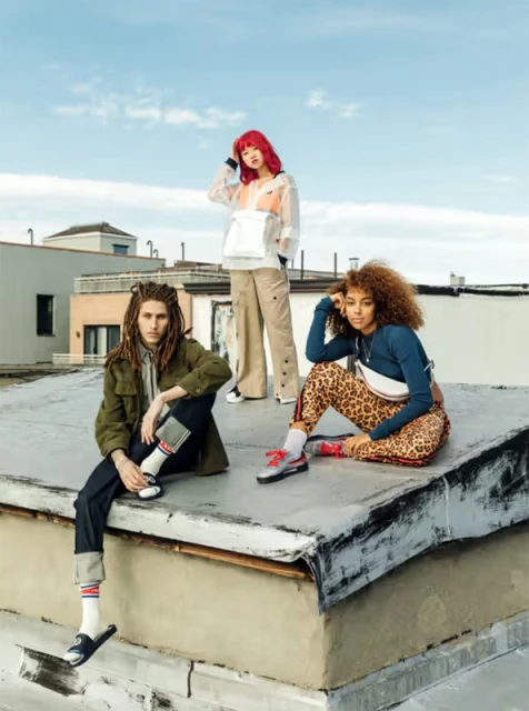 Three fashion models pose on a rooftop wearing stylish casual clothing, including jackets, pants, sneakers, and socks, against a blue sky.