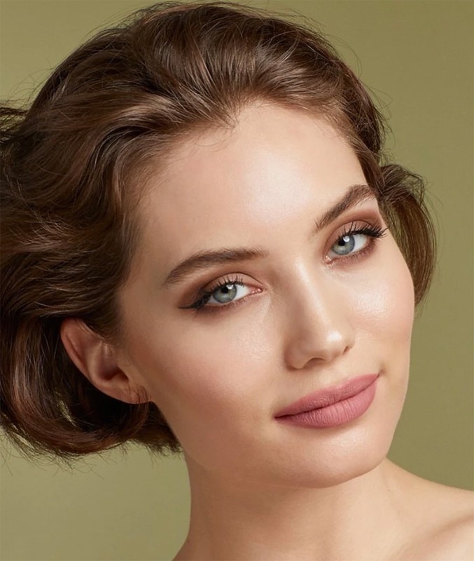 Close-up portrait of a woman with wavy brown hair, wearing makeup and looking at the camera against a muted green backdrop.