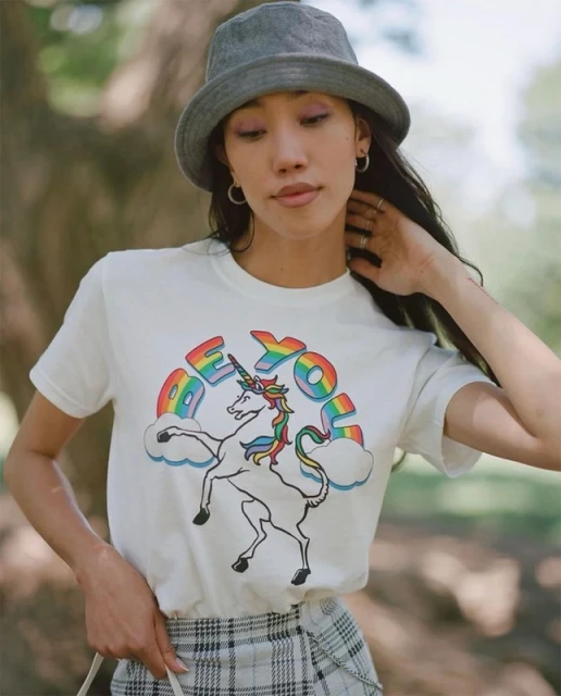 An Asian woman is wearing a t-shirt with unicorn graphics and a gray bucket hat in an outdoor setting with natural light.