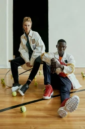 Two models seated on a polished wooden indoor court surrounded by tennis balls with hair styled by Janera Rose