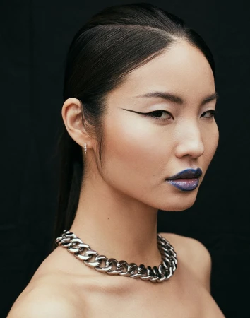 Close-up portrait of an East Asian woman with dramatic winged eyeliner, metallic blue lips, and a chunky silver chain necklace.