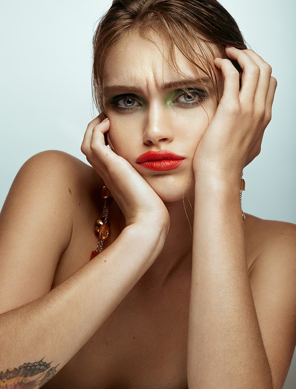 Close-up portrait of a woman with orange lipstick, green eyeshadow, and wet hair, posing with her hands on her face in a photo studio.