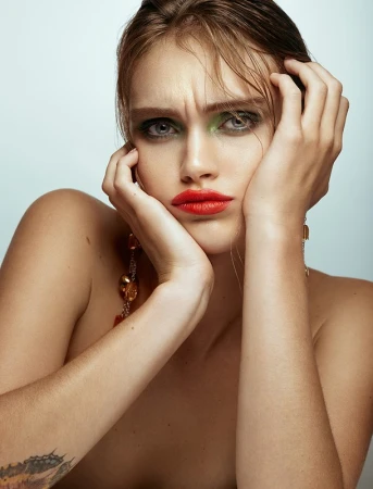 Close-up portrait of a model with green eyeshadow and bright red lipstick, holding hands to face against light grey