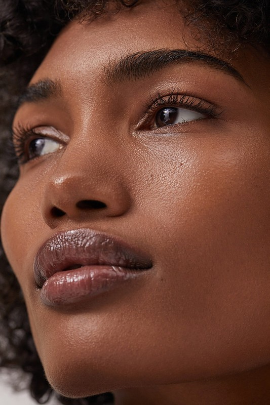 Close-up portrait of a woman with dark curly hair and glossy lips, showcasing her skin's texture and makeup in a neutral indoor setting.