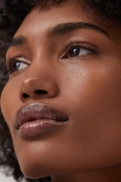 Close-up portrait of a woman with dark curly hair and glossy lips, showcasing her skin's texture and makeup in a neutral indoor setting.