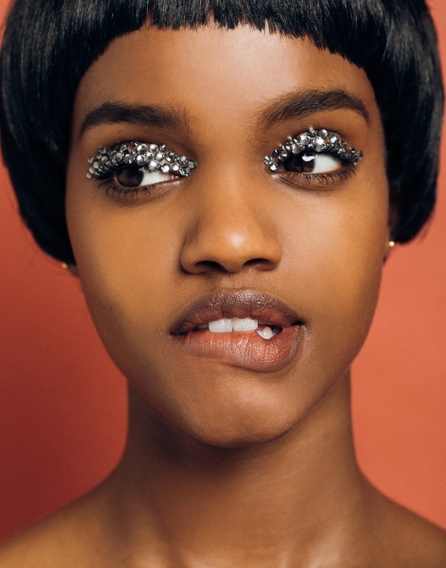 Close-up portrait of a black woman with glitter eyeshadow and dark hair biting her lip against a reddish-brown backdrop.