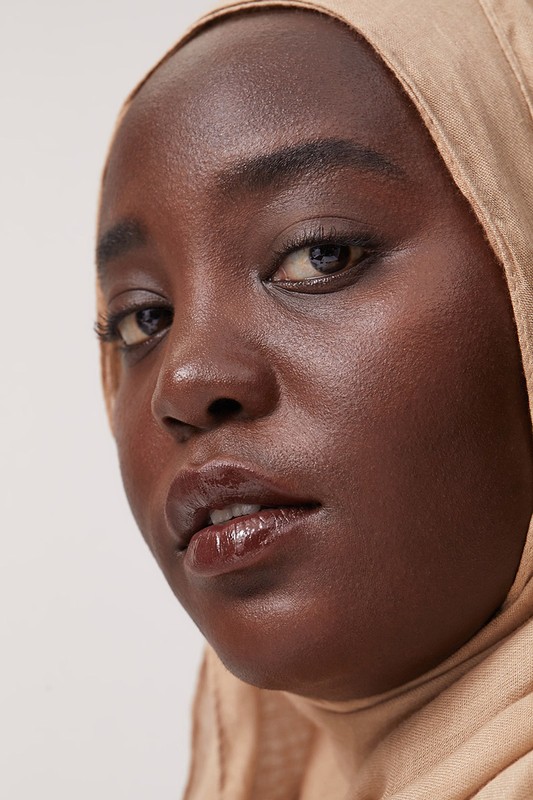 Close-up portrait of a Black woman wearing a beige hijab and glossy lip makeup against a neutral background.