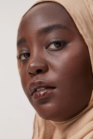 Close-up portrait of a Black woman wearing a beige hijab and glossy lip makeup against a neutral background.