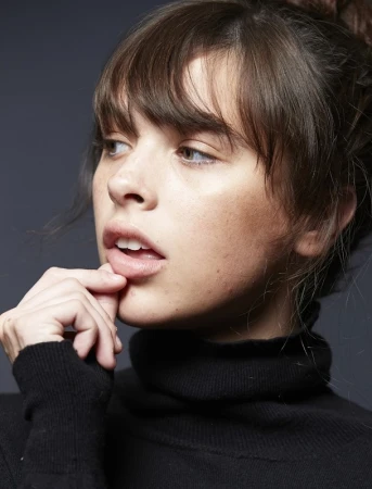 Close-up portrait of a woman with bangs wearing a black turtleneck and touching her lips
