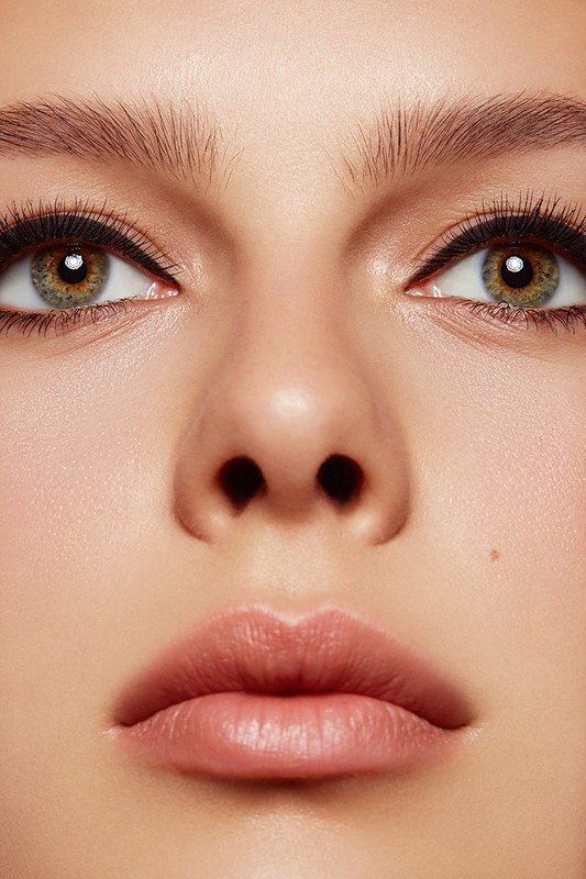 Close-up portrait of a woman's face featuring hazel eyes, light pink lips, and fair skin with natural makeup in a soft, diffused light.